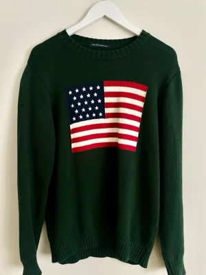 Mörkgrön stickad tröja Brandy Melville - Mörkgrön stickad tröja från Brandy Melville med amerikansk flagga i rött, vitt och blått på bröstet. Klassisk rund halsringning och långa ärmar. Perfekt till kyligare dagar för en chill och snygg preppig, amerikansk look. I nytt skick, enbart testad en gång. 