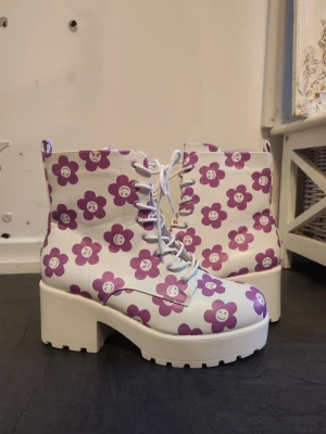 Koi boots med lila blommor - Säljer ett par chunky boots från Koi med vit bas och lila blommönster med olika ansikten. Skorna har snörning, plattformsula och hög klack. Materialet är syntet och de har rund tå. Perfekt för dig som vill sticka ut med färgglada detaljer. Använda fåtal gånger