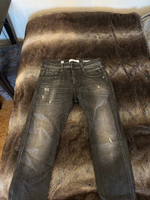 Replay grå jeans med slitningar - Säljer ett par grå jeans från Replay med snygga slitningar och tvättade detaljer. Modellen har klassisk femficksdesign, normal midja och raka ben. Jeansen är tillverkade i bomull och har en cool, lite sliten look som passar perfekt till streetwear.