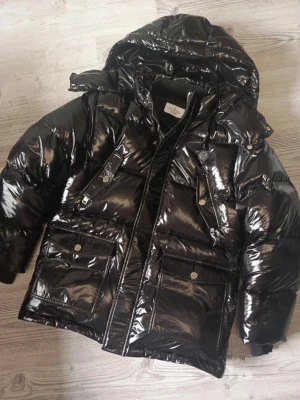 Svart pufferjacka från Plein - Säljer en svart, glansig pufferjacka från Plein med huva och feta fickor framtill. Jackan har en cool döskalle på ryggen och dragkedja med döskalle-detalj. Materialet är syntet och polyester, perfekt för kalla dagar. Grym streetstil och riktigt schysst look.