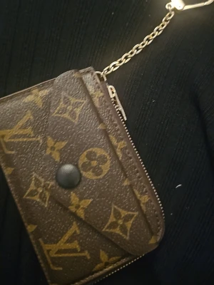 brun plånbok med kedja - Säljer en brun plånbok med klassiskt monogrammönster i guld. Plånboken har en praktisk kedja med karbinhake, flera kortfack på baksidan och dragkedja runt kanten. Materialet är skinn och detaljerna är i guldton. Perfekt för kort och småsaker.