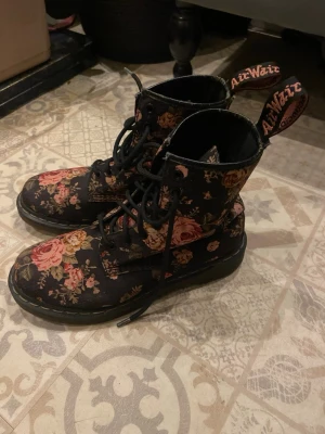 Svarta Dr. Martens boots med blommigt mönster - Säljer ett par svarta Dr. Martens boots med snyggt blommigt mönster. Skorna har rund tå, snörning och klassisk AirWair-detalj på hälen. Perfekt för dig som vill sticka ut med din stil.