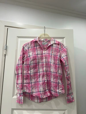 Vintage skjorta från Morris Lady - Säljer en vintage rutig skjorta från Morris Lady i storlek 38. Skjortan har en snygg mix av rosa, vit, blå och orange i rutmönster, klassisk krage och knappar framtill. Materialet är mjuk bomull och det finns en liten broderad detalj på bröstet.