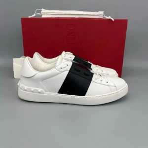 Valentino Garavani Open sneakers vit/svart - Säljer ett par Valentino Garavani Open sneakers i vitt skinn med bred svart rem över mitten. Skorna har rund tå, platt sula och vita nitar på hälen för extra edge. Klassisk snörning och logga på plösen. Perfekt för dig som gillar stilrena designer.