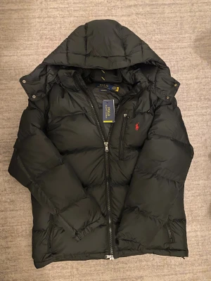 Ralph Lauren Jacka - Svart pufferjacka från Polo Ralph Lauren med huva, tvåvägsdragkedja och ikonisk röd logga på bröstet. Jackan har quiltad design och flera fickor med dragkedja. Perfekt för kalla dagar och har en clean, sportig look.