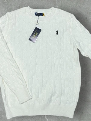 Vit stickad tröja från Polo Ralph Lauren - Säljer en vit stickad tröja från Polo Ralph Lauren med klassiskt kabelstickat mönster och broderad logga på bröstet. Tröjan har rund hals och långa ärmar, perfekt för dig som gillar stilrena och tidlösa plagg.