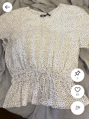 Vit prickig blus från Zara - Säljer en söt vit blus från Zara med svarta prickar. Blusen har korta ärmar, rund hals och en volang nedtill. Midjan är markerad med smock och knytband för extra detalj. Perfekt att styla med jeans eller kjol för en fräsch look.