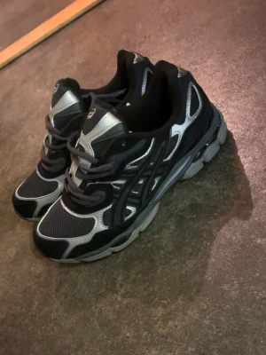 Svarta och silver Asics sneakers - Säljer ett par Asics sneakers i svart och silver med mesh och syntetmaterial. Skorna har snörning, rund tå och platt sula. Asics GEL-dämpning för extra komfort och cool design med kontrasterande detaljer. Perfekt för dig som gillar sportig stil. Vi kan diskutera priset.