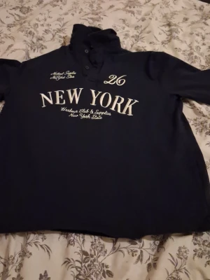 Mörkblå henley med New York-tryck - Säljer en mörkblå henley med vit broderad text 'NEW YORK' och siffran 26 på bröstet. Skjortan har knappar vid halsen och lång ärm. Materialet är mjuk bomull och passformen är normal. Perfekt för dig som gillar sportig och urban stil.