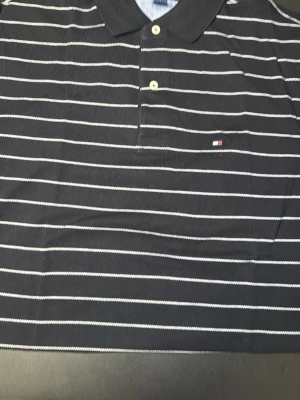 Randig pikétröja från Tommy Hilfiger - Säljer en svart pikétröja från Tommy Hilfiger med vita horisontella ränder och klassisk krage. Tröjan har knappar framtill och den ikoniska lilla loggan broderad på bröstet. Perfekt till jeans eller shorts för en clean look.