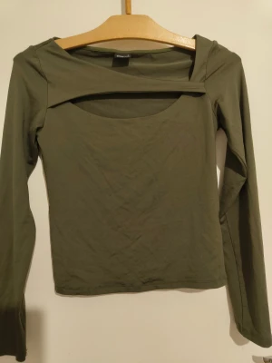 Fest topp Gina Tricot - Snygg olivgrön långärmad topp från Gina Tricot i storlek S. Du kan antingen ha den till en fest eller bara ut med vänner, jättefin och mjuk. Toppen har en unik skärning med öppen detalj vid halsen och är tillverkad i mjuk bomull. Perfekt för dig som gillar stilrena och trendiga plagg med en twist.
