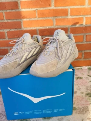 Adidas Ozelia sneakers ljusgrå 43 1/3 - Adidas Ozelia sneakers i ljusgrå färg med chunky siluett och retro vibes. Skorna har snörning, Adiprene-dämpning och detaljer i syntet och mocka. Ikonisk Adidas-logga på plösen och coola paneler på sidorna. Perfekt för dig som gillar streetwear och bekväma sneakers.