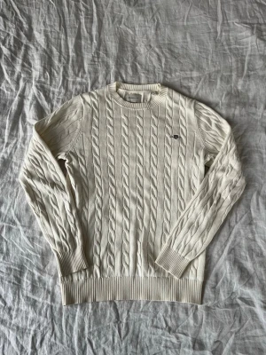 Vit kabelstickad tröja från GANT - Säljer en kräm vit kabelstickad tröja från GANT i storlek S. Tröjan har rund hals, ribbade muddar och ett litet broderat GANT-emblem på bröstet. Materialet är mjuk bomull och modellen är klassisk med lång ärm och normal passform.