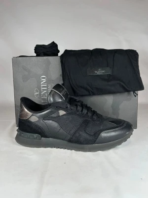 Valentino Garavani Rockrunner sneakers - Priset är ej hugget i sten!!                              Snygga svarta Valentino Garavani Rockrunner sneakers med detaljer i mocka och skinn. Skon har en rund tå, platt sula och coola nitar på hälen. Camouflage-mönster på vissa paneler ger en extra edge. 