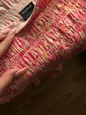 Rosa kortklänning från Gina Tricot - Säljer en söt kortklänning från Gina Tricot i storlek S. Klänningen har ett smockat liv och volang nedtill, med ett rosa och ljusgult mönster som ger en fräsch och somrig vibe. Materialet är mjuk bomull och passformen är otrolig och sitter jätte bra💕