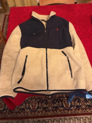 Beige teddyjacka från Polo Ralph Lauren - Säljer en beige teddyjacka från Polo Ralph Lauren med mörkblå detaljer och bröstficka med logga. Jackan har dragkedjor på fickorna och framtill, samt hög krage. Perfekt för kyliga dagar och riktigt skön att ha på sig.