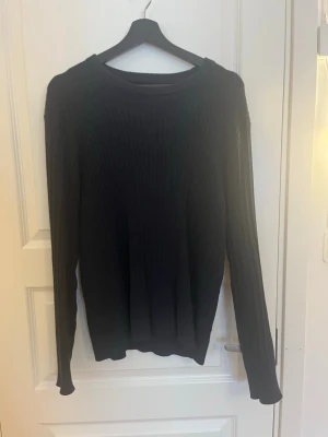 Svart ribbad stickad tröja från brothers  - Säljer en mörkblå stickad tröja med ribbad struktur och rund hals. Tröjan har lång ärm och en klassisk, enkel design som funkar till många olika stilar. Perfekt för kyliga dagar och enkel att matcha med jeans eller chinos. 