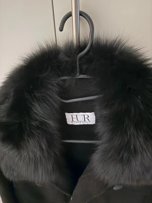 Svart pälsjacka från FUR Stockholm - Säljer en svart pälsjacka från FUR Stockholm med fluffig krage i päls och snygga knappar framtill. Jackan har två stora fickor med lock och en loose passform som ger en cool vibe. Perfekt för dig som vill sticka ut med stil. Storlek M passar nog 36/38.