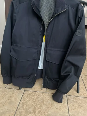 Svart vindjacka från J.Lindeberg - Svart vindjacka från J.Lindeberg med stilren design, stora fickor framtill och ribbade muddar. Jackan har dragkedja och en diskret logotyp på ärmen. Perfekt för dig som gillar minimalistisk och funktionell stil.