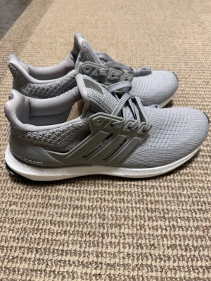Grå Adidas Ultraboost sneakers - Säljer ett par grå Adidas Ultraboost sneakers med stickad ovandel och klassiska tre ränder på sidan. Skorna har snörning, platt sula och en bekväm Boost-mellansula. Perfekta för dig som gillar stilrena och sportiga sneakers med modern design.