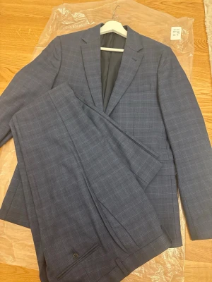 Blå rutig kostym från Riley Slim Fit - Säljer en stilren blå kostym från Riley i slim fit-modell. Kavajen och byxorna har ett diskret rutmönster och klassiska detaljer som knappar och fickor. Kostymen är fodrad med mörkgrått tyg och har en modern passform som ger en snygg siluett. Använd bara en gång aldrig mer köptes slutet av sommaren      St på byxor 50 på kavaj 48