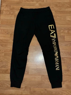 Svarta mjukisbyxor EA7 Emporio Armani - Svarta mjukisbyxor från EA7 Emporio Armani med guldtryck längs ena benet. Byxorna har resår i midjan och vid bensluten, samt sidofickor. Perfekta för chill eller träning, med en riktigt snygg och modern look.