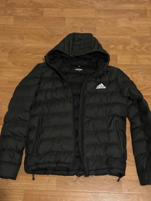 Svart pufferjacka från Adidas - Säljer en svart pufferjacka från Adidas med huva och dragkedja framtill. Jackan har ett stilrent, quiltat mönster och Adidas-loggan i vitt på bröstet. Perfekt för kyliga dagar och enkel att matcha med olika outfits.