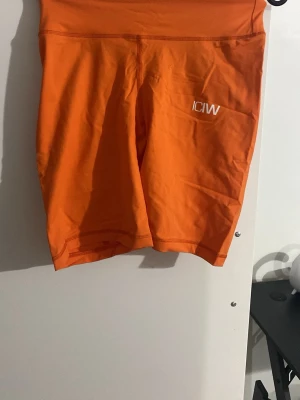 Orange scrunch biker shorts från ICANIWILL - Säljer ett par orange scrunch V-shape biker shorts från ICANIWILL. De har hög midja, logga framtill och scrunch-effekt bak. Materialet är stretchigt med SWEATTECH-teknologi, tillverkat av 77% nylon och 23% spandex. Perfekta för träning och riktigt snygga på gymmet.
