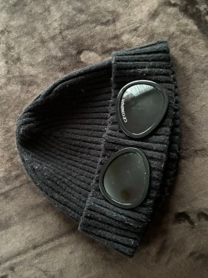 Svart mössa från C.P. Company - Svart ribbstickad mössa från C.P. Company med två ikoniska svarta glasögon-linser framtill. Mössan har en cool och urban vibe, perfekt för dig som vill sticka ut. Materialet är mjukt och stretchigt för skön passform.