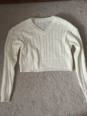 Vit croppad stickad tröja från SHEIN - Säljer en vit croppad stickad tröja från SHEIN i storlek XS. Tröjan har ett snyggt flätmönster, v-ringad hals och långa ärmar. Perfekt för dig som gillar en chill och trendig stil. Ribbstickade muddar vid ärmslut och nederkant.