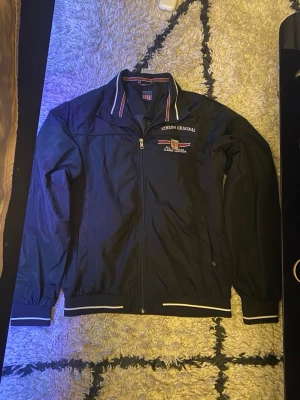 Svart vindjacka från Vinson Original - Svart vindjacka från Vinson Original med broderad logga på bröstet och snygga ränder i kragen. Jackan har dragkedja framtill, ribbade muddar med vita detaljer och är tillverkad i polyester. Perfekt för dig som gillar sportig och clean stil.