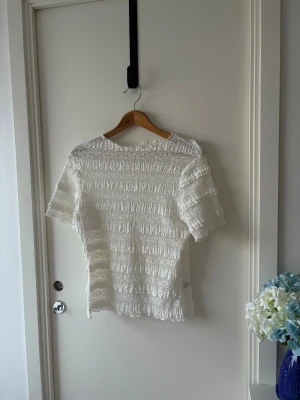Vit blus med spets och volanger - Säljer en vit blus med korta ärmar, täckt av spets och volanger i horisontella rader. Blusen har en rund hals och är tillverkad i ett lätt, luftigt material som ger en romantisk och feminin känsla. Perfekt för dig som gillar detaljer och en söt stil. Står ingen storlek men är stretchig i material och passar mig väl som vanligtvis bär M/L