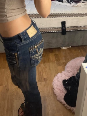 Blå bootcut jeans med broderi och dragkedjor - Säljer ett par mörkblå jeans från RMC med bootcut passform. Jeansen har snygga guldfärgade dragkedjor och broderade detaljer på bakfickorna. Klassisk femficksmodell med låg midja och slitningar framtill för en cool look.