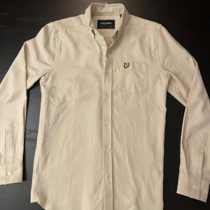 Beige skjorta från Lyle & Scott - Säljer en stilren beige skjorta från Lyle & Scott med klassisk krage och knappstängning hela vägen. Skjortan har en bröstficka med den ikoniska gula fågeln broderad.