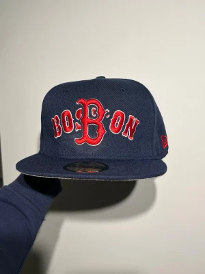 Boston  New Era 59FIFTY keps - Snygg mörkblå Boston Red Sox keps från New Era, modell 59FIFTY. Framtill står det 'BOSTON' i röd broderad text med vit kant och stor B-logga. På sidan finns New Era-loggan i rött och på baksidan står det 'BOSTON'. Undersidan av skärmen är ljusgrå.