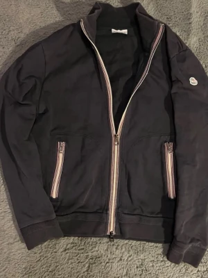 Svart zip-kofta från Moncler - Säljer en svart kofta från Moncler med hel dragkedja och två fickor med randiga detaljer. Tröjan har långärmad design och Moncler-logga på ena ärmen. Perfekt för dig som gillar stilrena och sportiga plagg.