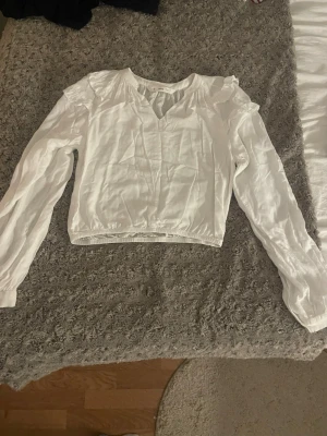 Vit blus från Mango- stl S - Vit, luftig blus från Mango med fina volangdetaljer vid axlarna och lätt puffärm. V-ringad framtill och resår i midjan som ger en smickrande passform. Långärmad och i tunt, mjukt material – perfekt till både vardag och finare tillfällen. (Kort modell) 🤍