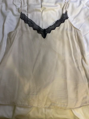 Vit  linnetopp med svart spets H&M - Säljer en vit linnetopp från H&M i storlek XS. Toppen har tunna axelband och en snygg v-ringning med svart spetsdetalj framtill. Materialet är lent och har en lätt glans, perfekt för att styla med jeans eller kjol.