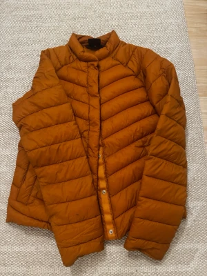 Orange pufferjacka i storlek 36 - Säljer en orange ”dun” jacka i storlek 36. Jackan har en quiltad design med tryckknappar framtill och står upp lite i kragen. Materialet är syntet och jackan är lätt vadderad, perfekt som mellanlager för kyligare dagar eller under höst/vår. Enkel och clean look som passar till det mesta. Liten fläck på ärmen. Går troligtvis att få bort, har ej testat.