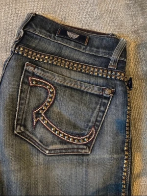Rock & Republic jeans med nitar - Snygga blå jeans från Rock & Republic med coola nitar längs fickor och sidor. Bakfickan har ett stort R med nitar och kontrastsömmar. Jeansen har klassisk femficksdesign och bootcut-ben. Perfekt för dig som gillar detaljer och vill sticka ut.