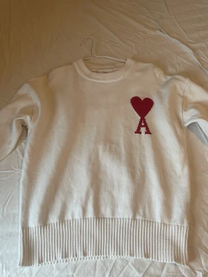 Vit stickad tröja med rött hjärta från Ami - Vit stickad tröja från Ami med ett rött hjärta och bokstaven A broderat på bröstet. Tröjan har rund hals, långa ärmar och bred ribbad kant nedtill och vid ärmslut. Perfekt för dig som gillar stilrena detaljer och vill ha något som sticker ut.