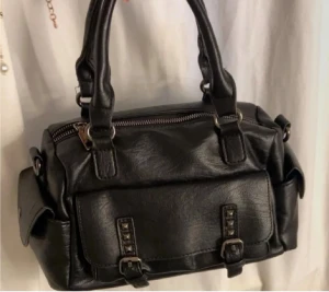 Svart handväska med nitar och dragkedja - Cool svart handväska i skinnimitation med två rejäla handtag och snygga nitar på framsidan. Väskan har praktiska fickor på sidorna och en stor dragkedja upptill. 