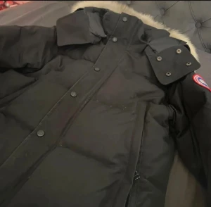 Canada Goose - Säljer en svart dunjacka från Canada Goose med stor fluffig päls på huvan. Jackan har dragkedja och knappar framtill, samt klassisk patch på ärmen. Perfekt för kalla vinterdagar och har en riktigt schysst oversized look.