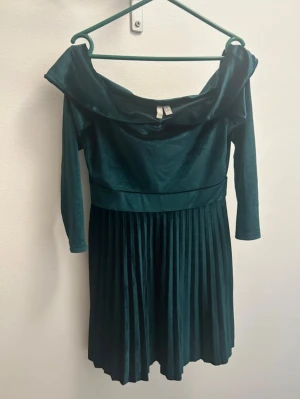 Mörkgrön plisserad sammetklänning - Supersnygg mörkgrön klänning från Asos i mjuk sammet med plisserad kjol och offshoulder-ringning. Klänningen har trekvartsärmar och markerad midja som ger en elegant siluett. Perfekt för dig som vill sticka ut med stil på nästa tillställning.