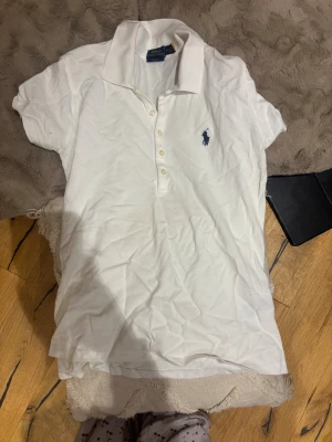 Vit pikétröja från Ralph Lauren - Vit pikétröja från Ralph Lauren i slim fit-modell. Klassisk krage, korta ärmar och knäppning med fyra knappar framtill. Ikoniska blå Ralph Lauren-loggan broderad på bröstet. Perfekt för en clean och sportig stil. I storlek medium men skulle säga att den passar dem i storlek s också