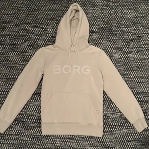 Beige hoodie från Björn Borg - Snygg beige hoodie från Björn Borg med stor BORG-logga på bröstet. Tröjan har huva med snörning, magficka och ribbade muddar vid ärmar och nederkant. Perfekt för chill eller träning, och tillverkad i mjuk bomull för skön känsla. PYTTELITEN MISSFÄRGNING PÅ FRAMSIDA SE BILD3. 