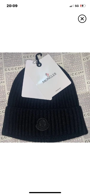 Svart ribbad mössa från Moncler - Svart ribbstickad mössa från Moncler med uppvikt kant och diskret Moncler-logga framtill. Snygg och enkel design som passar till allt, tillverkad i mjukt material som håller dig varm. Perfekt för dig som gillar stilrena accessoarer.