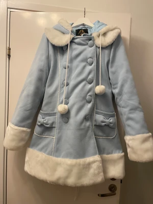 Ljusblå kappa från Hell Bunny - Superfin ljusblå pälsjacka från Hell Bunny med fluffig vit fuskpäls på krage, ärmslut och nederkant. Jackan har huva, snörning i ryggen, stora knappar framtill, rosetter på fickorna och söta pompoms. Quiltad insida för extra värme och mysig vinterstil.