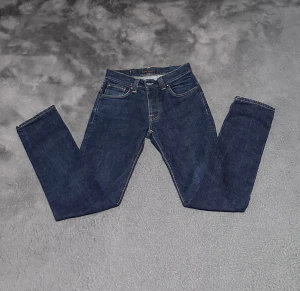 Mörkblå Nudie Jeans grim tim  - Snygga mörkblå jeans från Nudie Jeans med klassisk femficksdesign och kontrastsömmar. Modellen är Grim Tim och är tillverkad i slitstarkt jeanstyg av bomull. Bakfickorna har Nudie Jeans signaturbroderi och en brun läderpatch med logga. Riktigt bra skick som nya! Storlek 30/32 men känns mindre.
