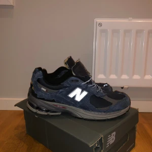 New Balance 2002R marinblå sneakers - Säljer ett par New Balance 2002R sneakers i marinblått med grå detaljer. Skorna har en chunky sula, rund tå och är tillverkade i mocka och syntet. Reflekterande N-logga på sidan och snörning framtill. Perfekta för dig som gillar streetwear och snygg komfort.